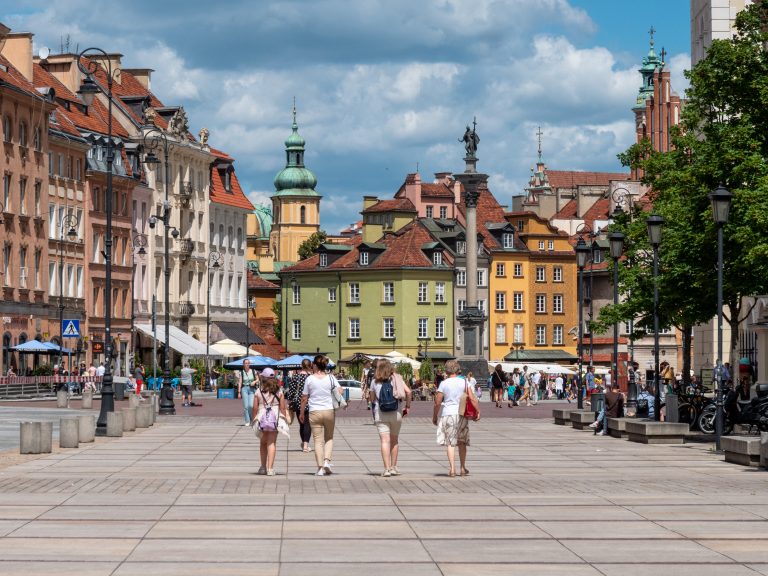 Zdjęcie przedstawia ulicę Krakowskie Przedmieście z widokiem na plac Zamkowy, w letni dzień. Na pierwszym planie widoczne cztery kobiety spacerujące w kierunku Kolumny Zygmunta III Wazy.