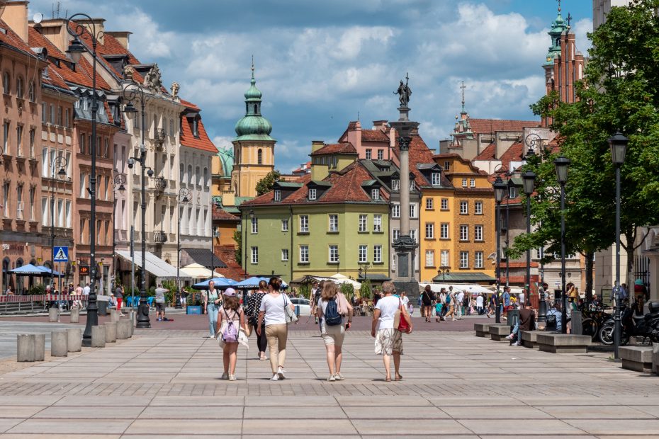 Zdjęcie przedstawia ulicę Krakowskie Przedmieście z widokiem na plac Zamkowy, w letni dzień. Na pierwszym planie widoczne cztery kobiety spacerujące w kierunku Kolumny Zygmunta III Wazy.