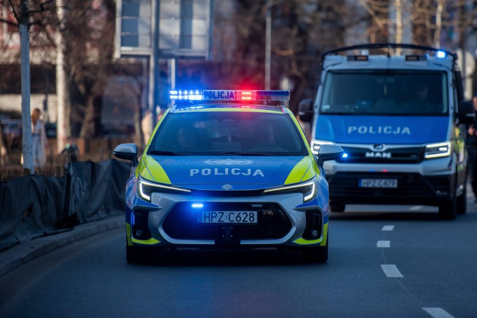 na pierwszym planie zdjęcia widoczny jest radiowozów policyjny z włączonymi sygnałami świetlnymi otwierający przejazd kolumny pojazdów
