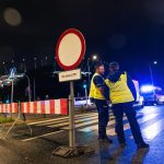 Zdjęcie przedstawia zaporę i znak drogowy B-1 zakaz ruchu w obu kierunkach ustawionych na wjeździe w aleję Zieleniecką. Obok znaku stoją policjanci pilnujący porządku w czasie wydarzenia na stadionie PGE Narodowym, który widoczny jest w tle.