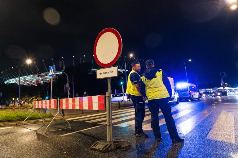 Zdjęcie przedstawia zaporę i znak drogowy B-1 zakaz ruchu w obu kierunkach ustawionych na wjeździe w aleję Zieleniecką. Obok znaku stoją policjanci pilnujący porządku w czasie wydarzenia na stadionie PGE Narodowym, który widoczny jest w tle.