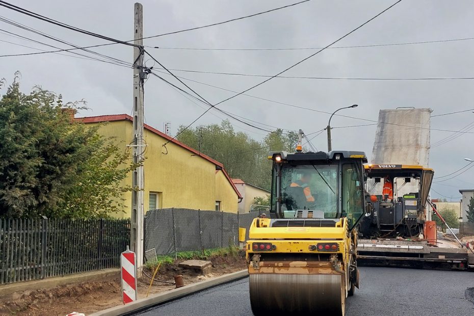 Zdjęcie przedstawia asfaltowanie nawierzchni niewielkiej ulicy. Na pierwszym planie widoczny walec drogowy, a za nim pracuje rozściełacz. Po lewej stronie widoczne rozkopane pobocze przygotowane do ułożenia chodnika. W tle domy jednorodzinne.