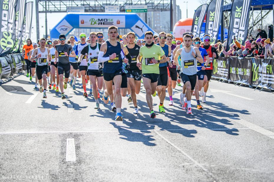 Fotografia przedstawia elitę biegaczy ruszających z pierwszej linii w zawodach BeMore5K. Zdjęcie wykonano chwilę po starcie. W tle widoczna jest brama startu i mety, a po prawej stronie kibice.