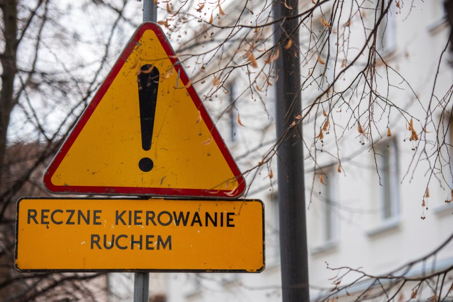 Zdjęcie przedstawia znak A-30 "inne niebezpieczeństwo" pod którym została umieszczona tabliczka z napisem "ręczne kierowanie ruchem". W tle widoczna kamienica, fragment latarni oraz gałęzie drzewa.