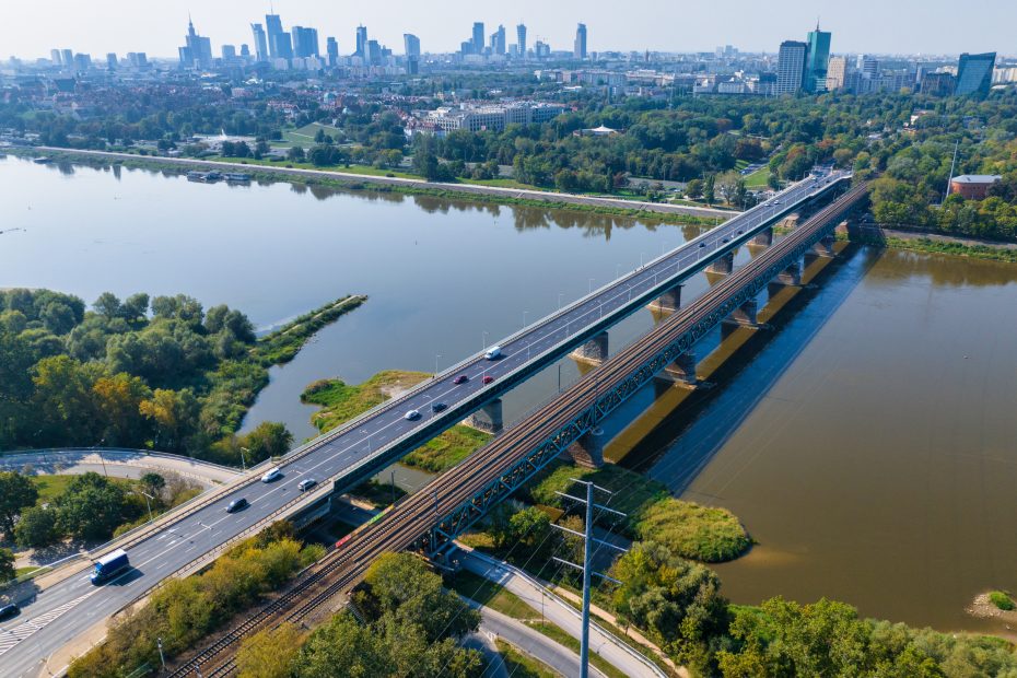 Fotografia przedstawia panoramę Warszawa, gdzie na pierwszym planie widoczny jest Most Gdański i sąsiadujący z nim most kolejowy. Zdjęcie wykonane w dzień, latem, widoczne dużo zieleni.