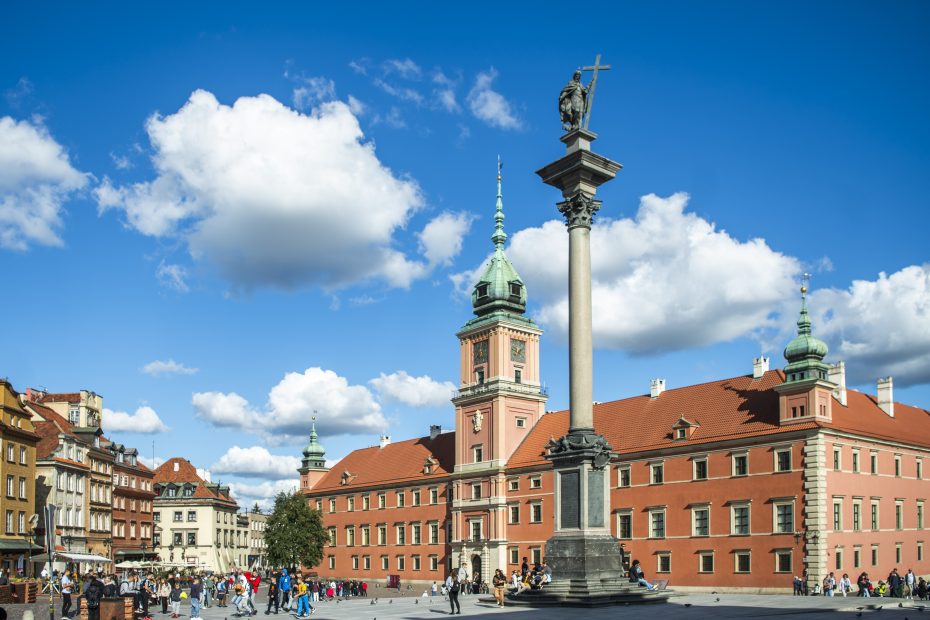 Zdjęcie przedstawia Plac Zamkowy w słoneczny dzień. Na placu widoczni tłumnie spacerujący spacerowicza. Na pierwszym planie widoczna jest Kolumna Króla Zygmunta III Wazy, a w tle Zamek Królewski.