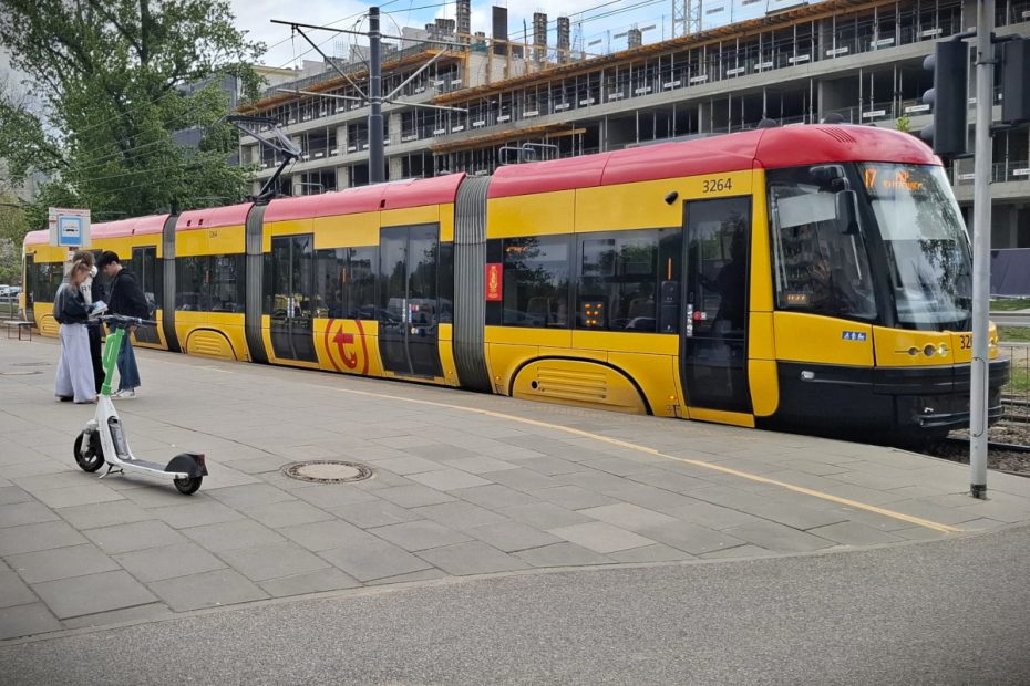 Zdjęcie przedstawia tramwaj stojący na przystanku Metro Słodowiec. Na pierwszym planie widoczna jest grupa młodych ludzi i hulajnoga.