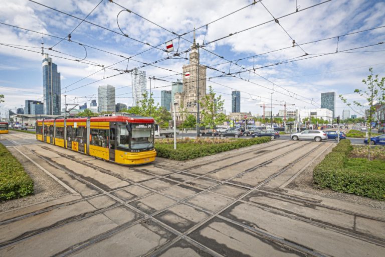 Zdjęcie przedstawia tramwaj przejeżdżający przez Rondo Dmowskiego. Na pierwszym planie widać krzyżujące się szyny i tramwaj, a w tle - Pałac Kultury i Nauki.