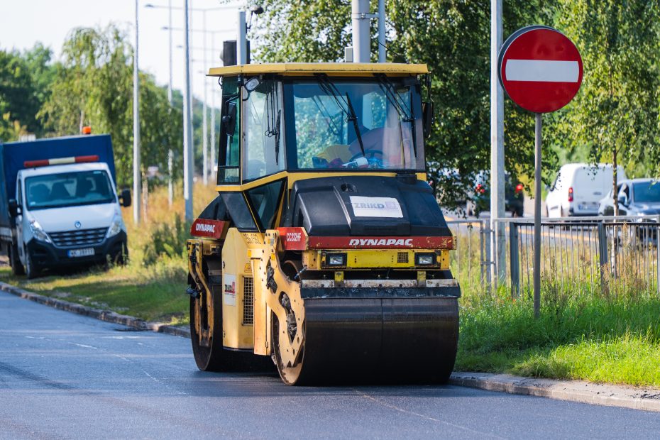 Zdjęcie przedstawia walec drogowy stojący na jedni. Obok niego widać znak zakaz wjazdu, a z tyłu ciężarówkę drogowców.