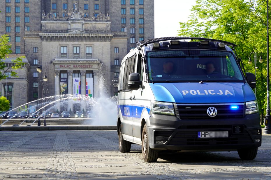 zdjęcie przedstawia duży radiowóz Policji stojący w Parku Świętokrzyskim na tle wejścia do Pałacu Młodzieży. W tle, z prawej strony widoczne są również drzewa, a na środku fontanna.