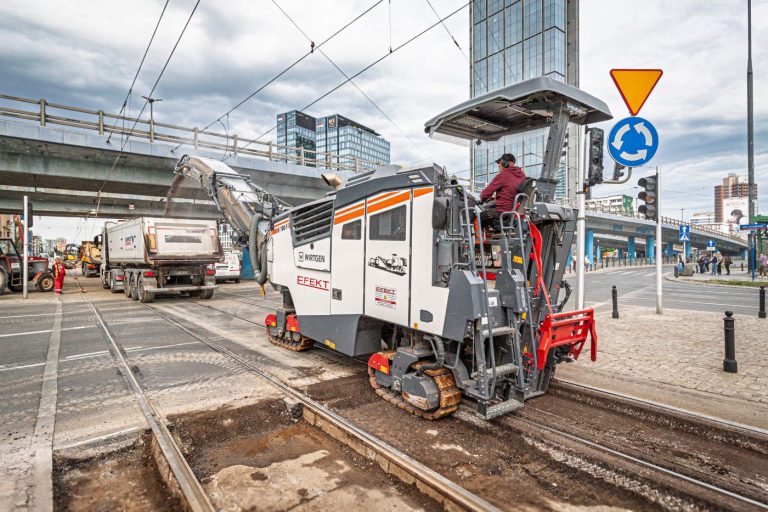 Zdjęcie przedstawia maszynę budowlaną pracującą na torowisku tramwajowym w Alejach Jerozolimskich przy Rondzie Czterdziestolatka.