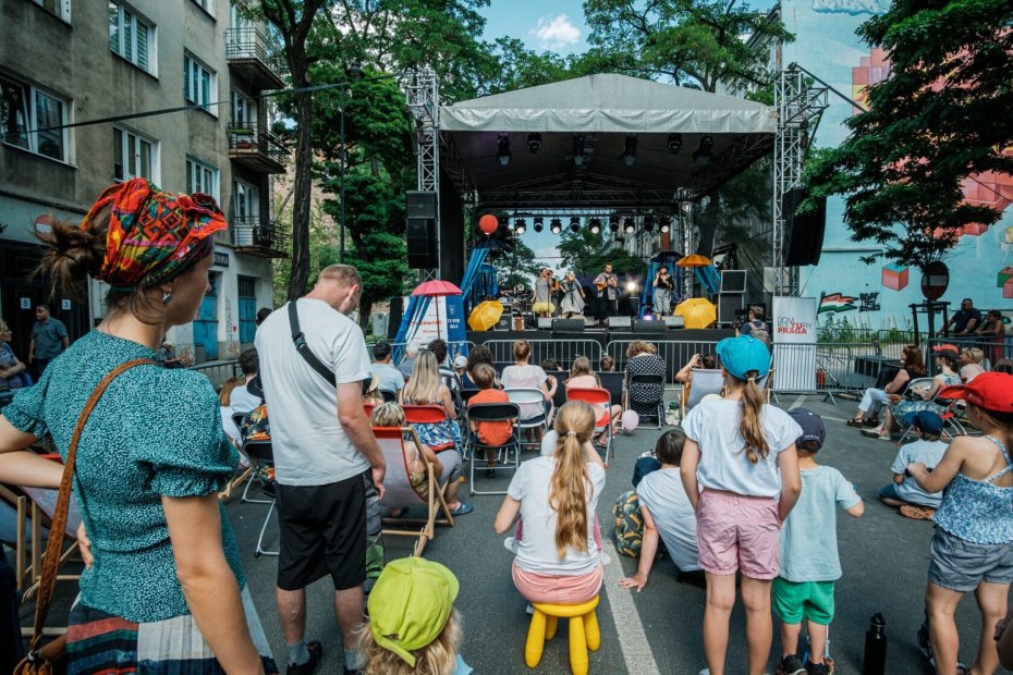Fotografia przedstawia koncert na scenie ustawionej na ulicy Stalowej w czasie festiwalu Święto ulicy Stalowej. Przed sceną widoczna grupa ludzi siedzących na różnego rodzaju krzesłach, a także bezpośrednio na jezdni. Z lewej strony zdjęcia widoczna jest kamienica, a po prawej fragment muralu.